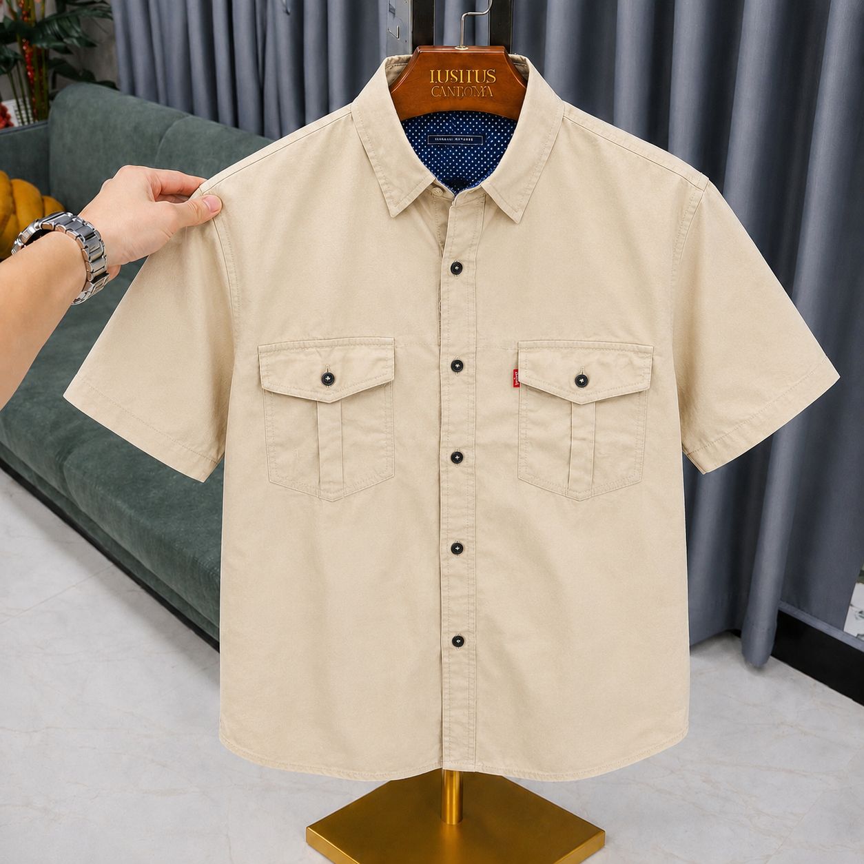 A beige short-sleeve button-up shirt with two chest flap pockets, hanging on a wooden hanger labeled JUSHITSU CLOTHING. A person's hand is holding the left shoulder of the shirt.