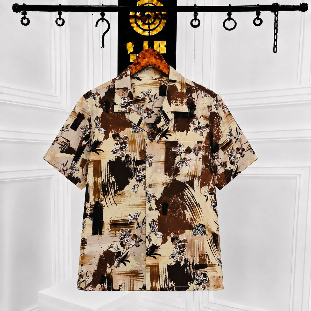A short-sleeve button-up shirt with an abstract floral print in beige, brown, and black, hanging on a wooden hanger against a white paneled wall.