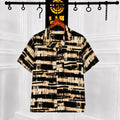 A short-sleeve button-up shirt with a black and beige abstract brushstroke pattern, hanging on a wooden hanger from a black clothing rack against a white paneled wall.