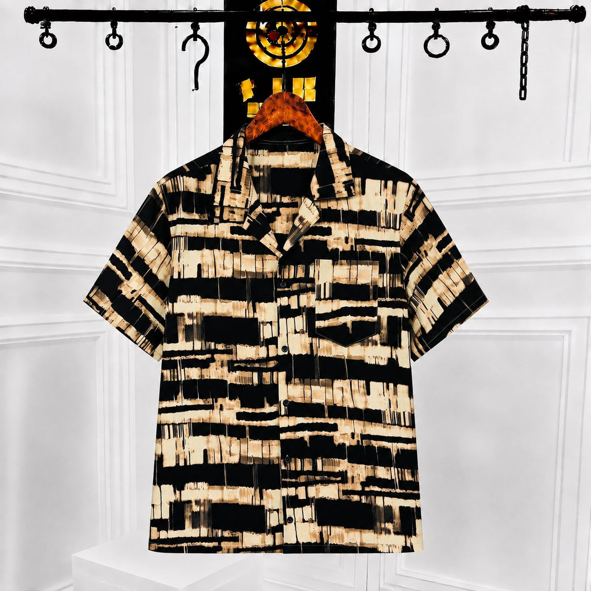 A short-sleeve button-up shirt with a black and beige abstract brushstroke pattern, hanging on a wooden hanger from a black clothing rack against a white paneled wall.