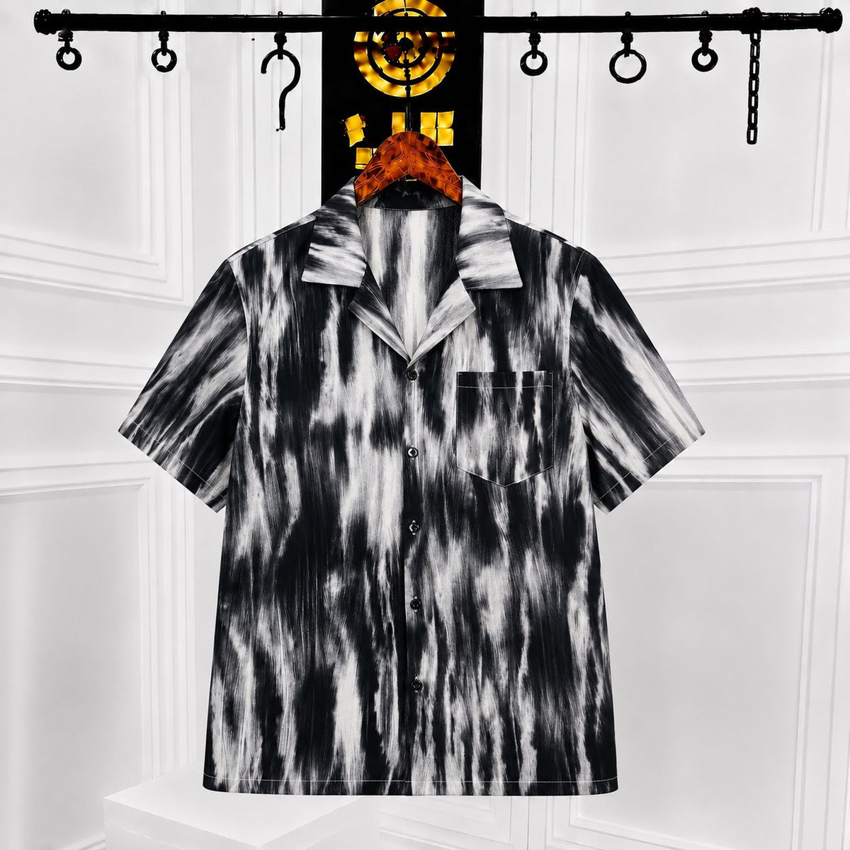 A short-sleeve button-up camp shirt with a black and white abstract tie-dye or brushstroke pattern, hanging on a hanger against a white paneled wall.