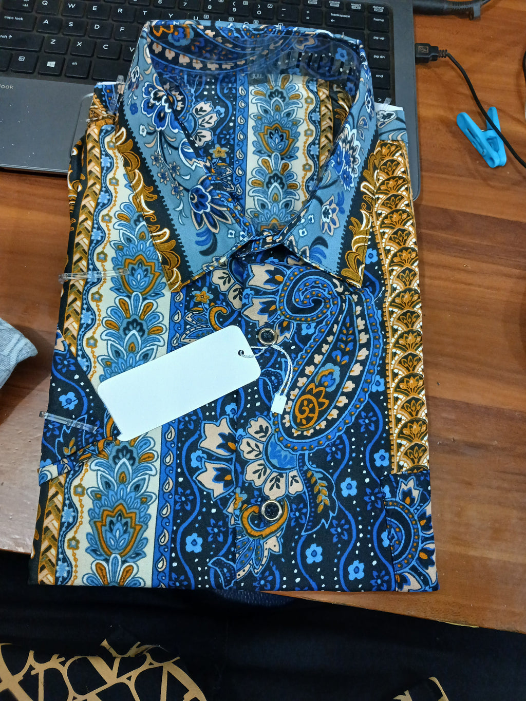 A folded blue and gold paisley printed shirt with a blank white tag, laid on a wooden desk next to a laptop keyboard.