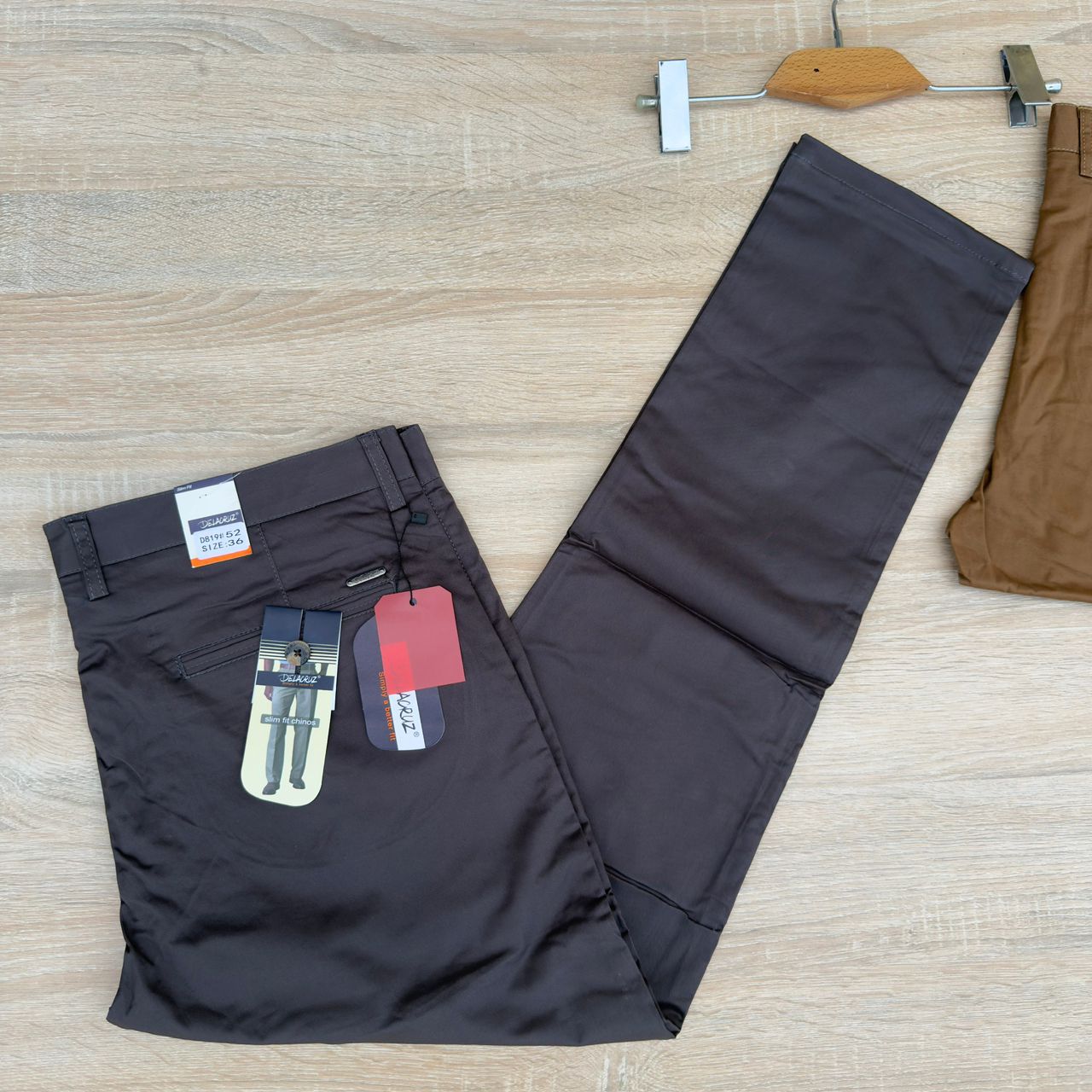 A pair of dark brown chino pants laid flat on a light wood floor, with retail tags attached and another brown garment partially visible at the right edge.