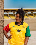 A woman wearing a yellow, red, and green short-sleeve jersey with a black star on the chest, standing in front of stadium seating.