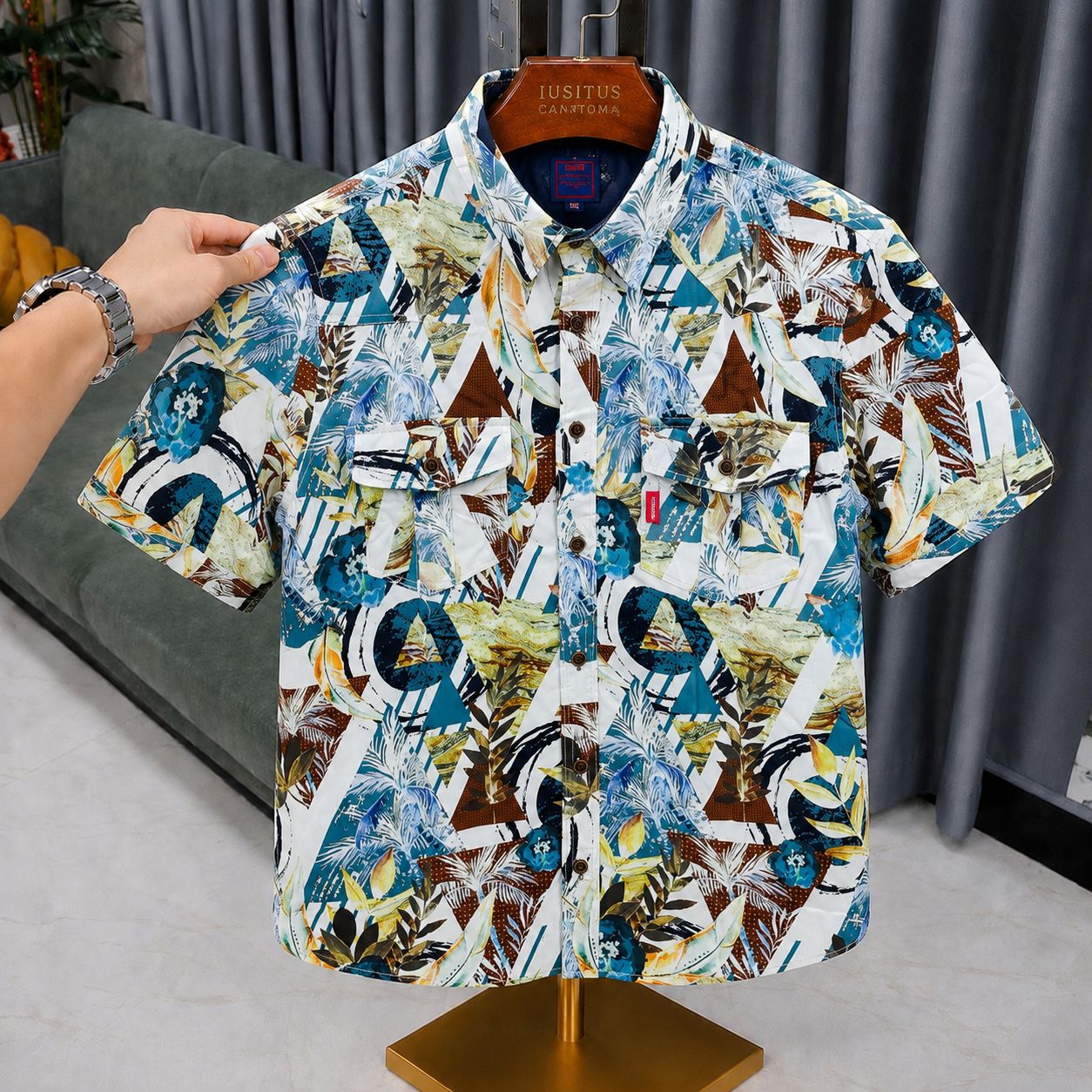 A short-sleeve button-up shirt with a colorful abstract geometric print, displayed on a wooden hanger labeled 'JUSSTERS CLOTHING' on a dress form stand. A hand is holding the left shoulder of the shirt.