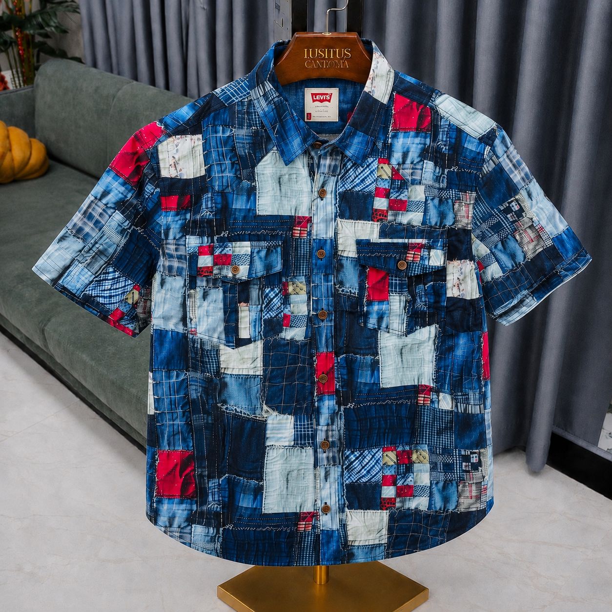 A short-sleeve button-up shirt with a colorful patchwork denim print, displayed on a wooden hanger labeled 'JUSLUS CLOTHING CO' on a mannequin stand. The background shows a gray sofa and curtains.