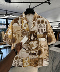 A men's short-sleeve button-up shirt with a brown and beige patchwork print featuring sunflowers, geometric shapes, and animal motifs, hanging on a black hanger in a clothing store. A hand is holding a white price tag in front of the shirt.