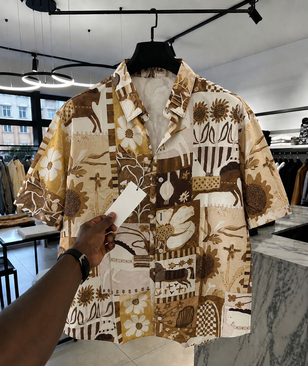 A men's short-sleeve button-up shirt with a brown and beige patchwork print featuring sunflowers, geometric shapes, and animal motifs, hanging on a black hanger in a clothing store. A hand is holding a white price tag in front of the shirt.