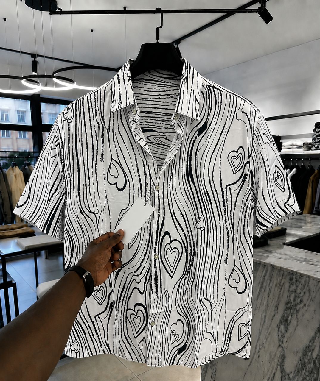 A black and white patterned short-sleeve button-up shirt with wavy lines and heart motifs, hanging on a black hanger in a retail store. A hand holding a white card is visible in the foreground.