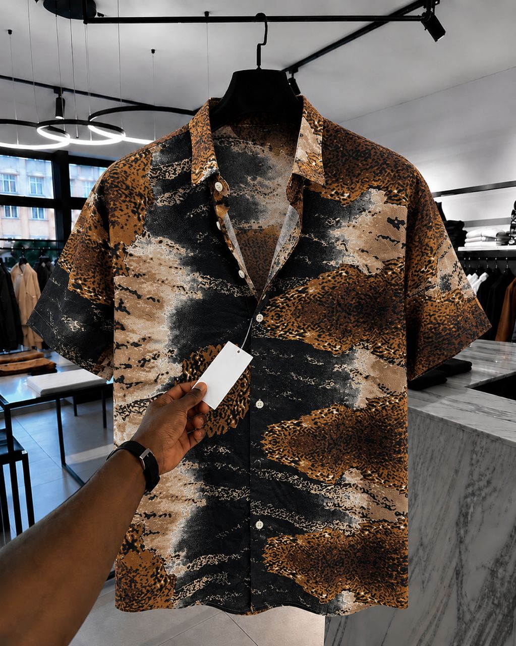 A short-sleeve button-up shirt with a brown, black, and beige abstract pattern, hanging on a black hanger in a clothing store. A hand is holding a white price tag attached to the collar.