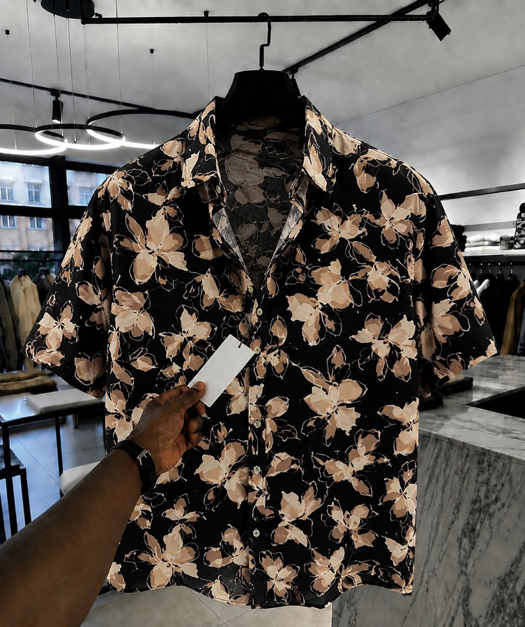 A short-sleeve button-up shirt with a black base and beige floral pattern, hanging on a black hanger in a retail store. A hand is holding a blank white price tag in front of the shirt.