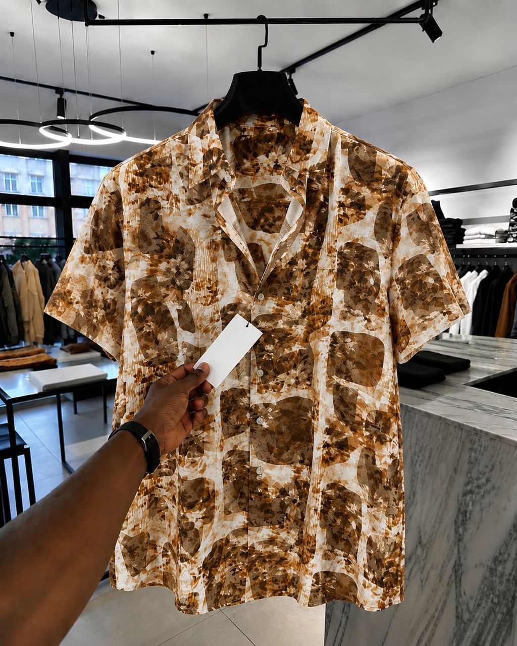 A man wearing a black watch holds up a short-sleeve button-up shirt with a brown and beige abstract pattern on a hanger inside a clothing store.