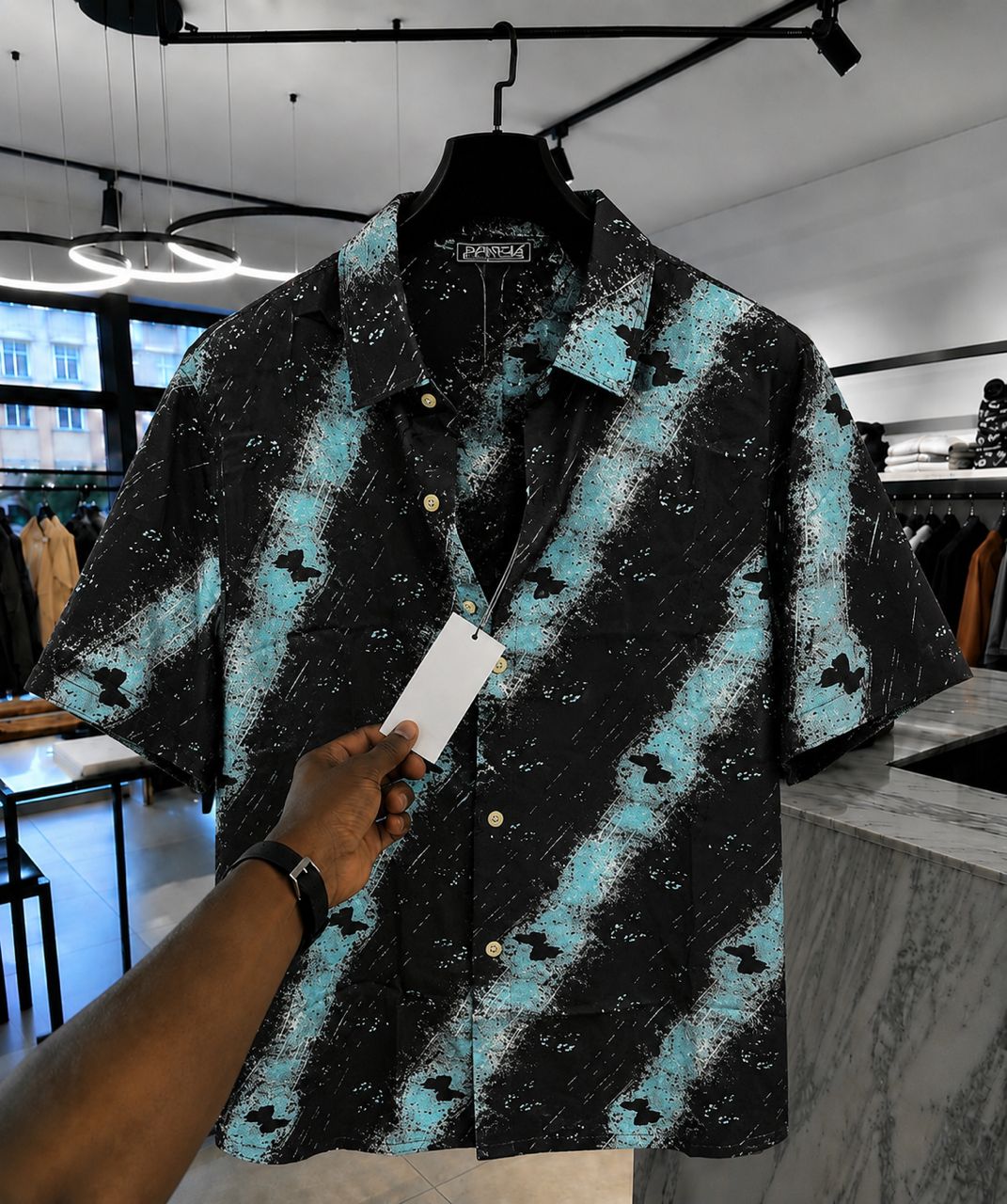 Black and blue patterned shirt on a hanger with a hand holding a card in a store setting.