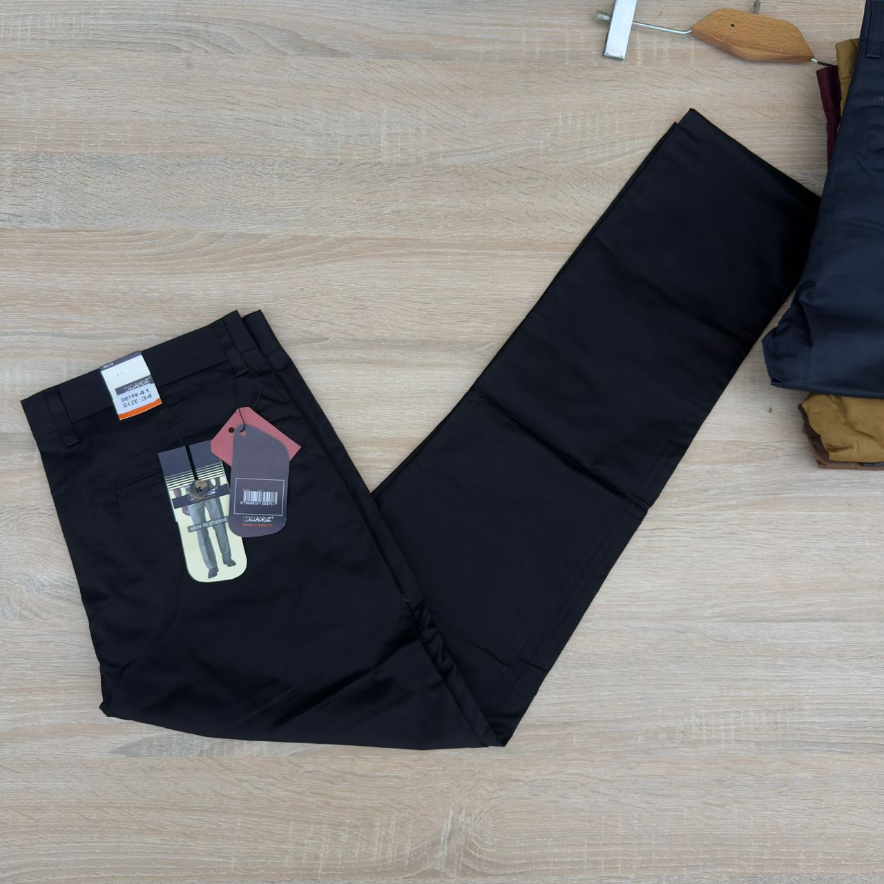 Black trousers laid flat on a light wood floor, with retail tags attached near the waistband and other folded clothing partially visible at the right edge.