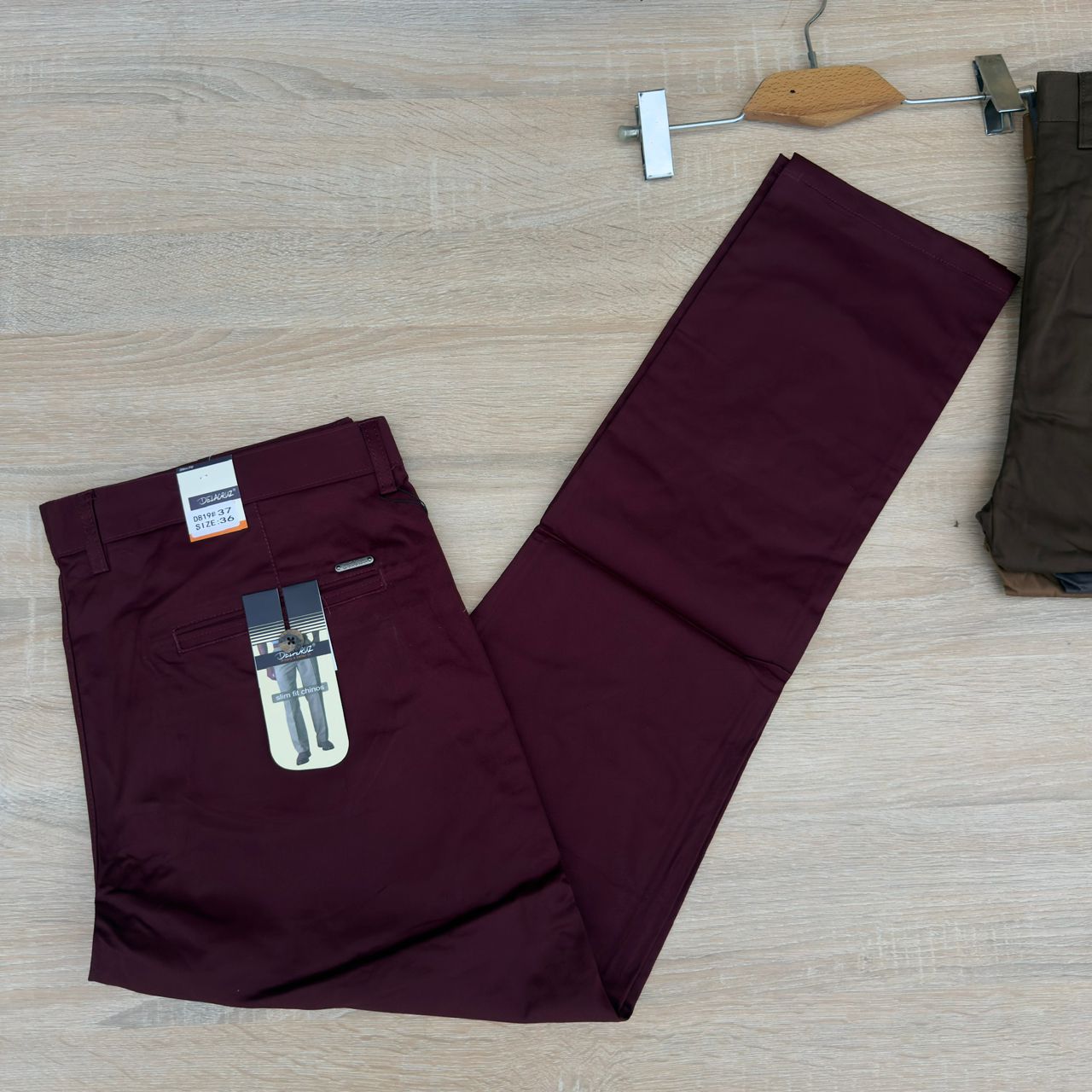 A pair of men's trousers in a deep burgundy/maroon color, laid flat on a light wood-grain floor. The pants are folded with a paper hangtag attached to the back pocket area.