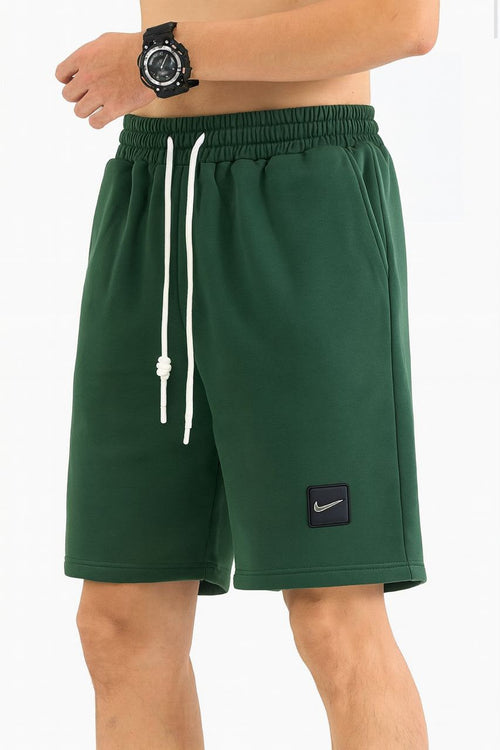 A person wearing dark green Nike athletic shorts with a white drawstring and a black Nike logo patch on the lower left leg, shown against a plain light background.
