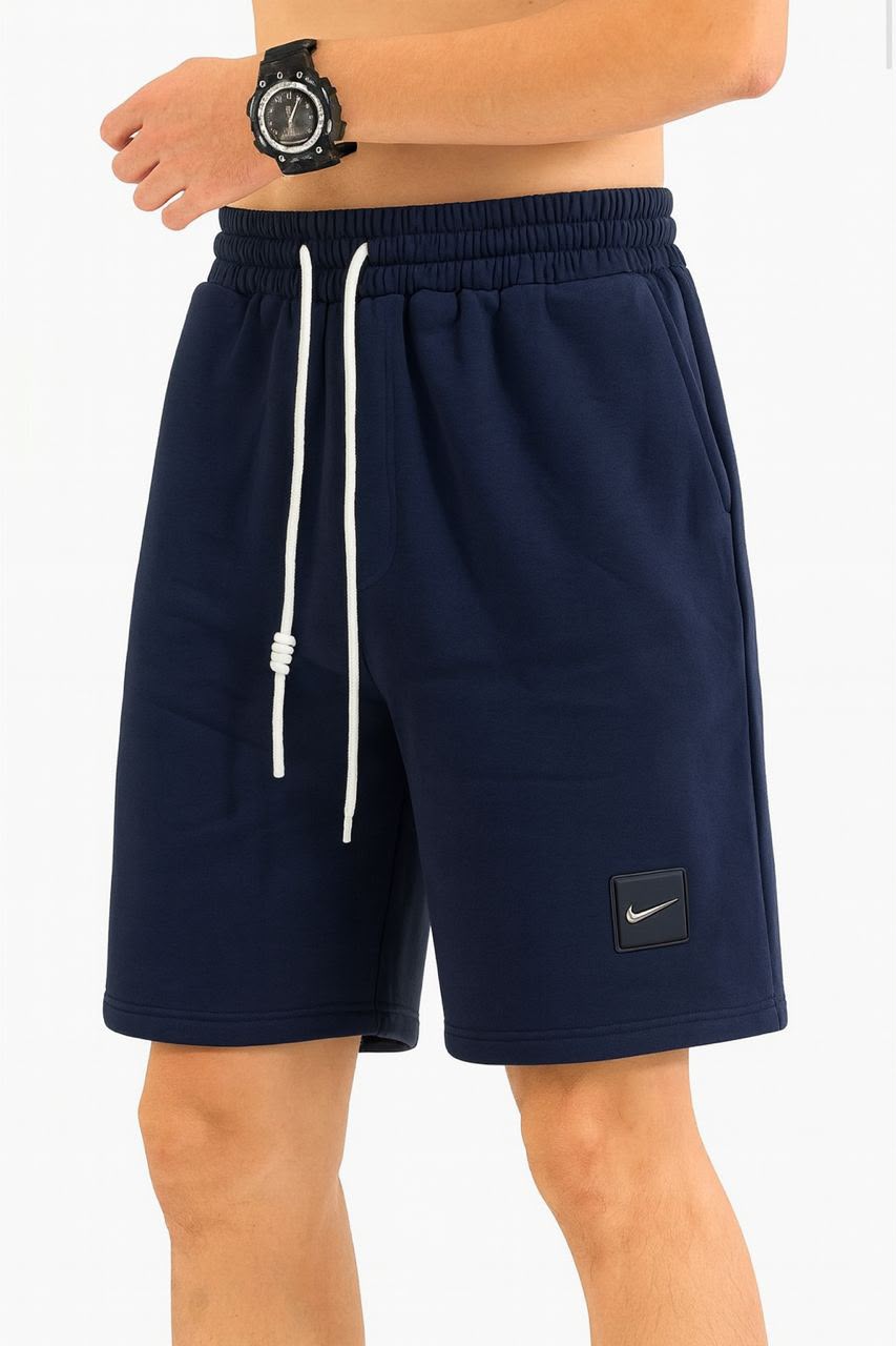 A person wearing navy blue Nike athletic shorts with a white drawstring and a small black Nike logo patch near the lower left leg, photographed against a light gray studio background.