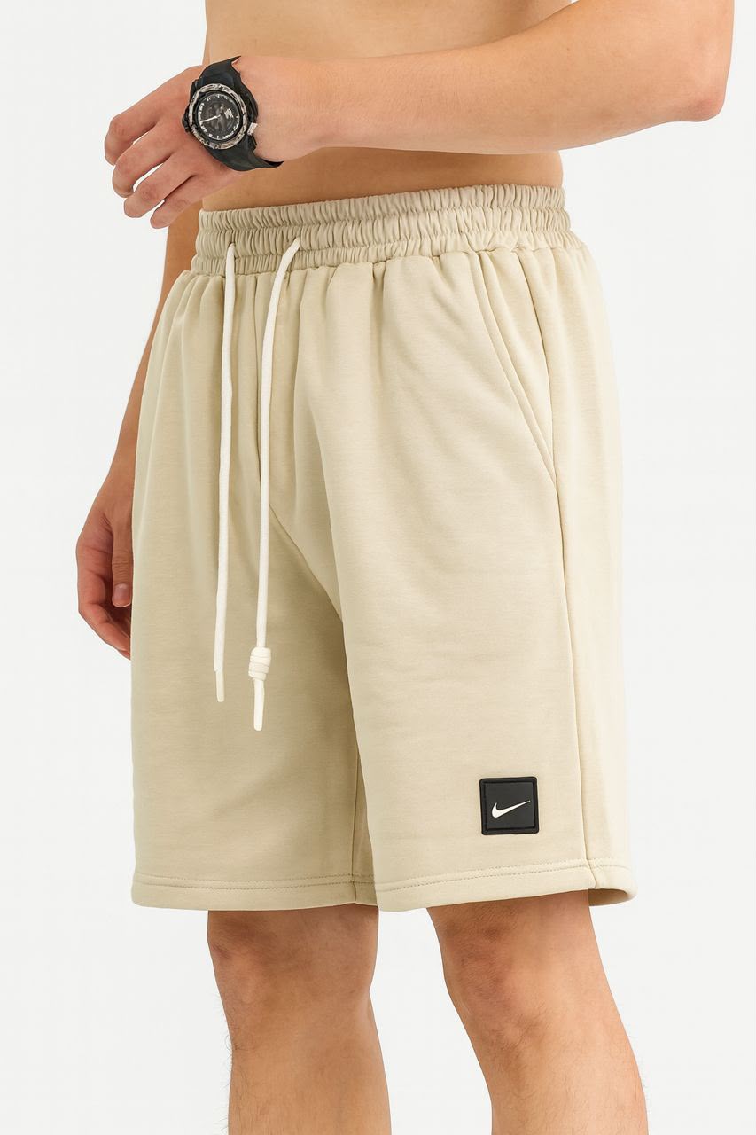 Beige athletic shorts with a white drawstring and a small black Nike logo patch on the lower left leg, shown on a model.