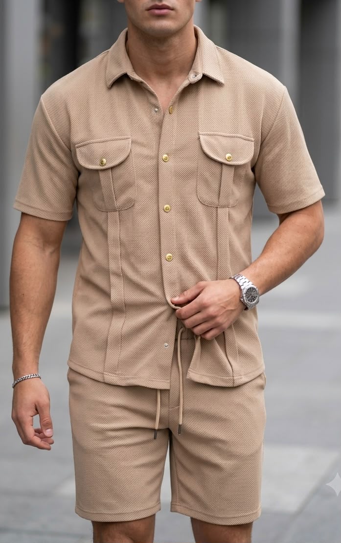A man wearing a matching beige short-sleeve button-up shirt and shorts set with gold-tone buttons, photographed outdoors against a blurred gray background.