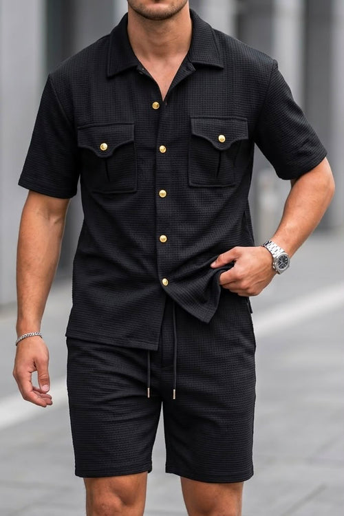 A man wearing a black short-sleeve button-up shirt and matching black shorts with gold-tone button accents, photographed outdoors on a paved walkway.