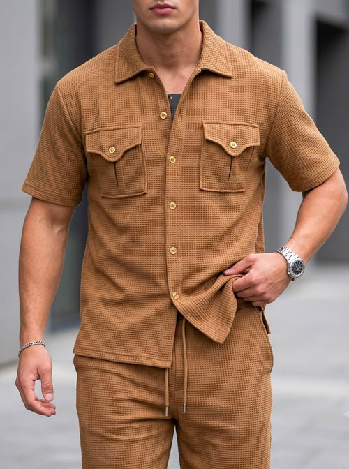 A man wearing a matching brown short-sleeve button-up shirt and drawstring shorts set, featuring a textured waffle pattern and gold-tone buttons, photographed outdoors against a blurred gray background.