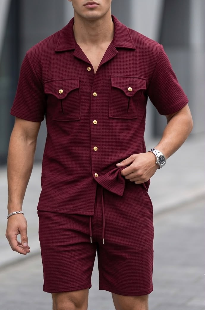 A man wearing a matching burgundy short-sleeve button-up shirt and shorts set with a subtle textured pattern and gold-tone buttons, photographed outdoors against a blurred gray background.