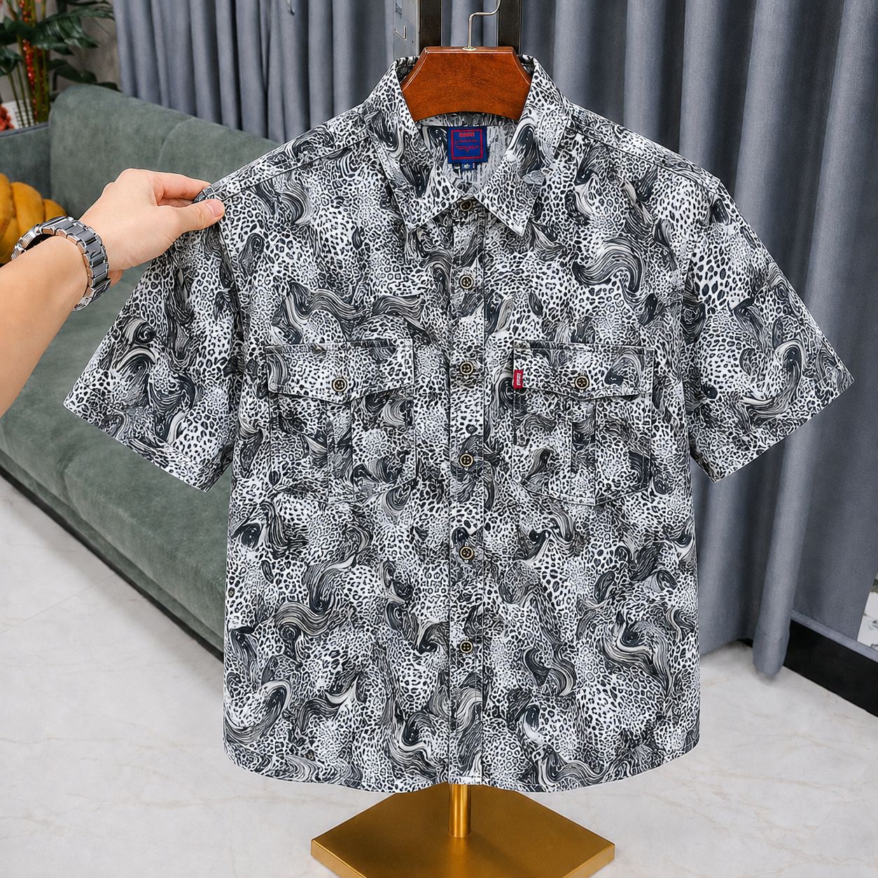 A short-sleeve button-up shirt with a black, white, and gray abstract pattern, displayed on a wooden hanger on a mannequin stand. A hand is holding the left shoulder of the shirt.