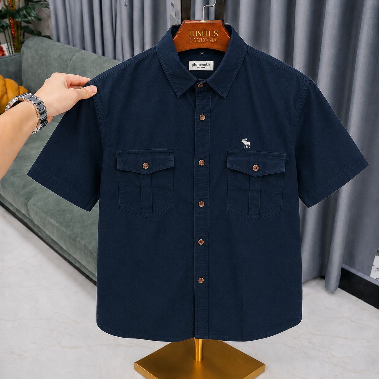 A dark navy blue short-sleeve button-up shirt with two chest pockets and a small white embroidered deer logo, displayed on a wooden hanger on a mannequin stand. A hand is holding the left shoulder of the shirt.