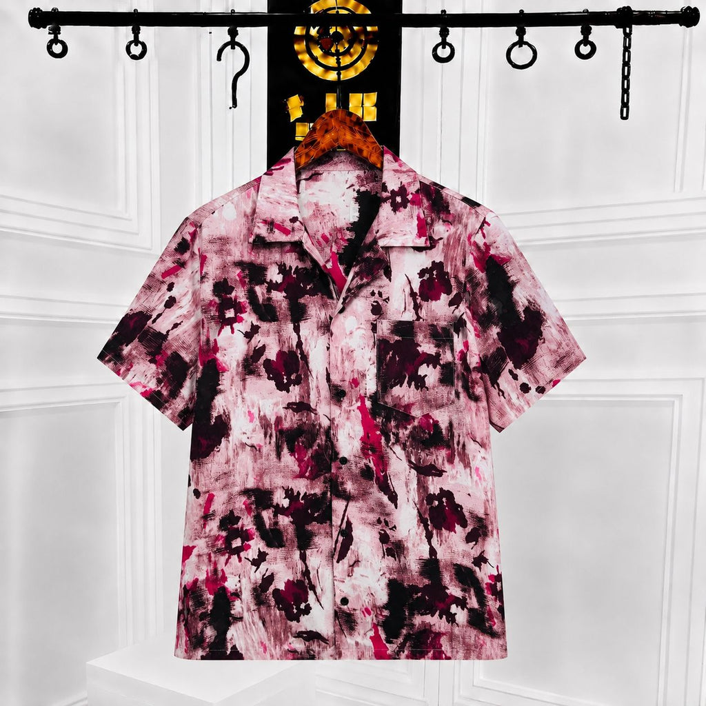 A short-sleeve button-up shirt with a pink, black, and white abstract paint-splatter pattern, hanging on a wooden hanger on a clothing rack against a white paneled wall.