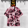 A short-sleeve button-up shirt with a pink, black, and white abstract paint-splatter pattern, hanging on a wooden hanger on a clothing rack against a white paneled wall.