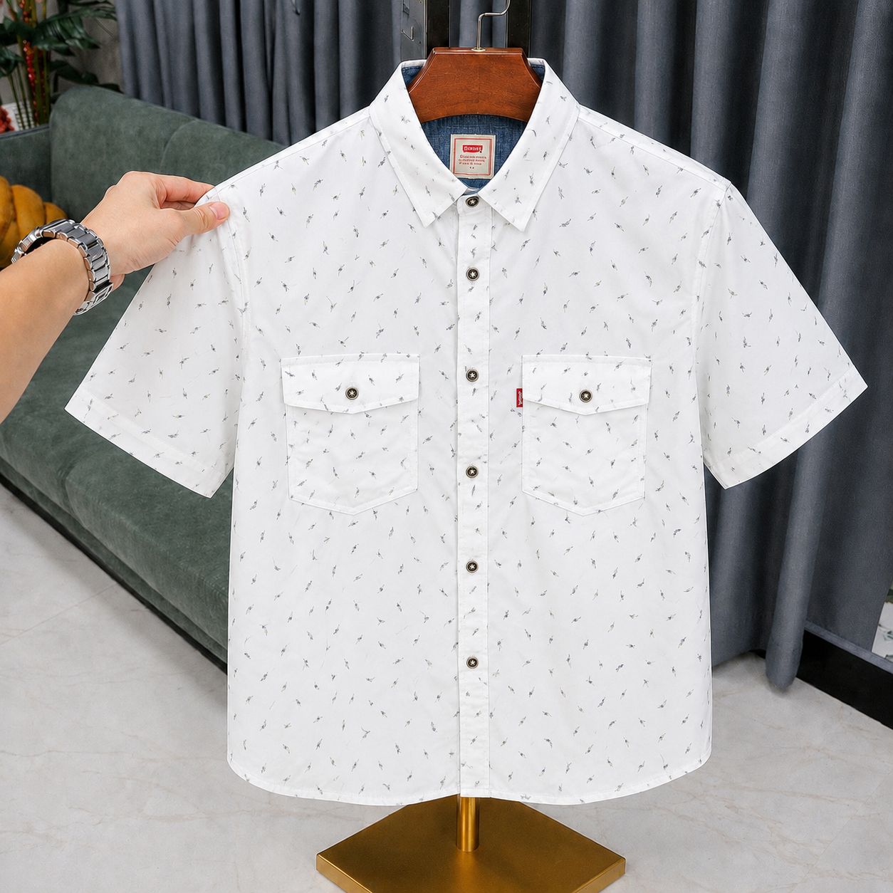 A white short-sleeve button-up shirt with a tiny all-over dark print, displayed on a hanger on a mannequin stand. A hand is holding the left shoulder of the shirt.