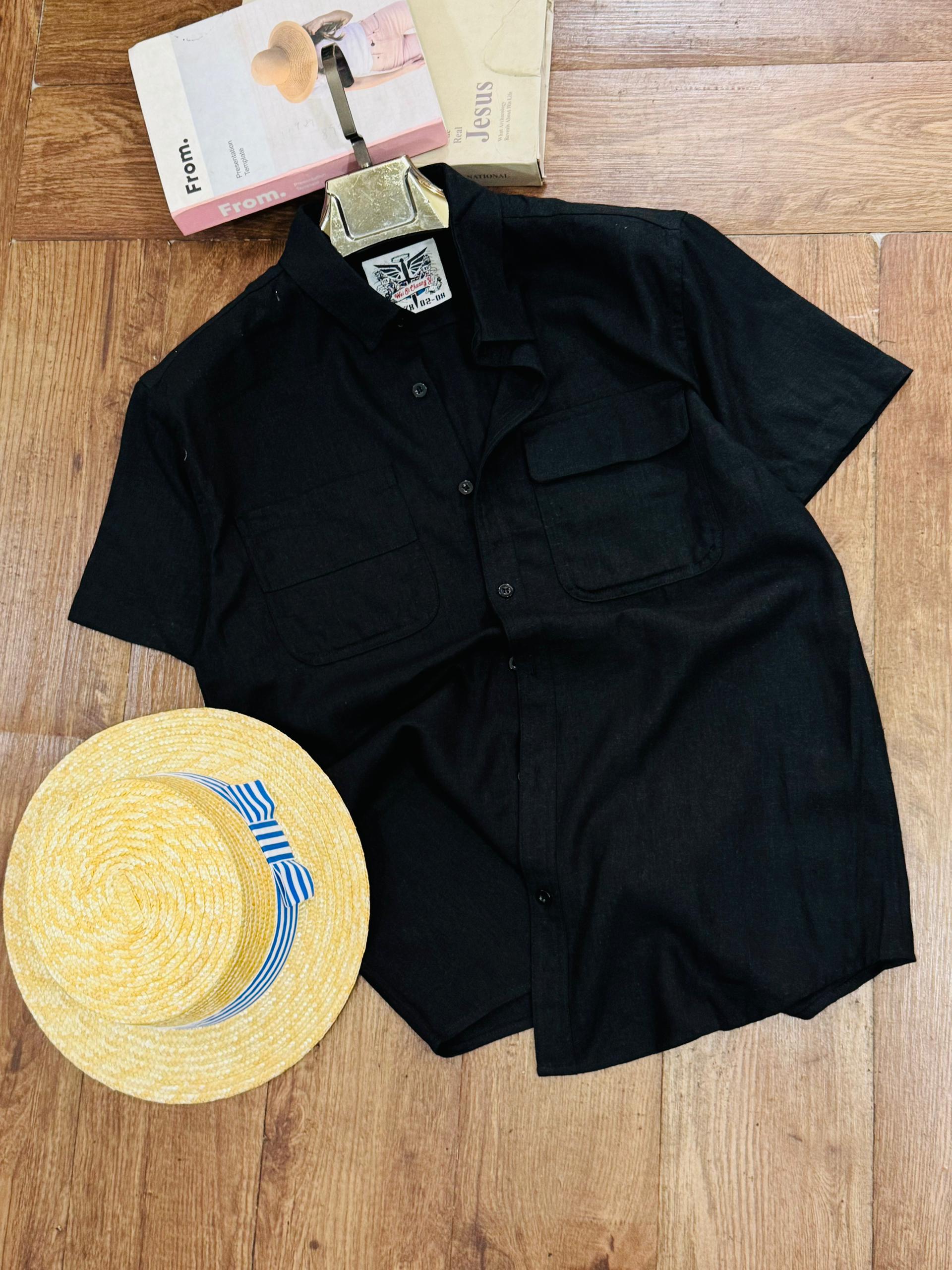A black short-sleeve button-up shirt laid flat on a wooden floor, styled with a yellow straw hat and a book.