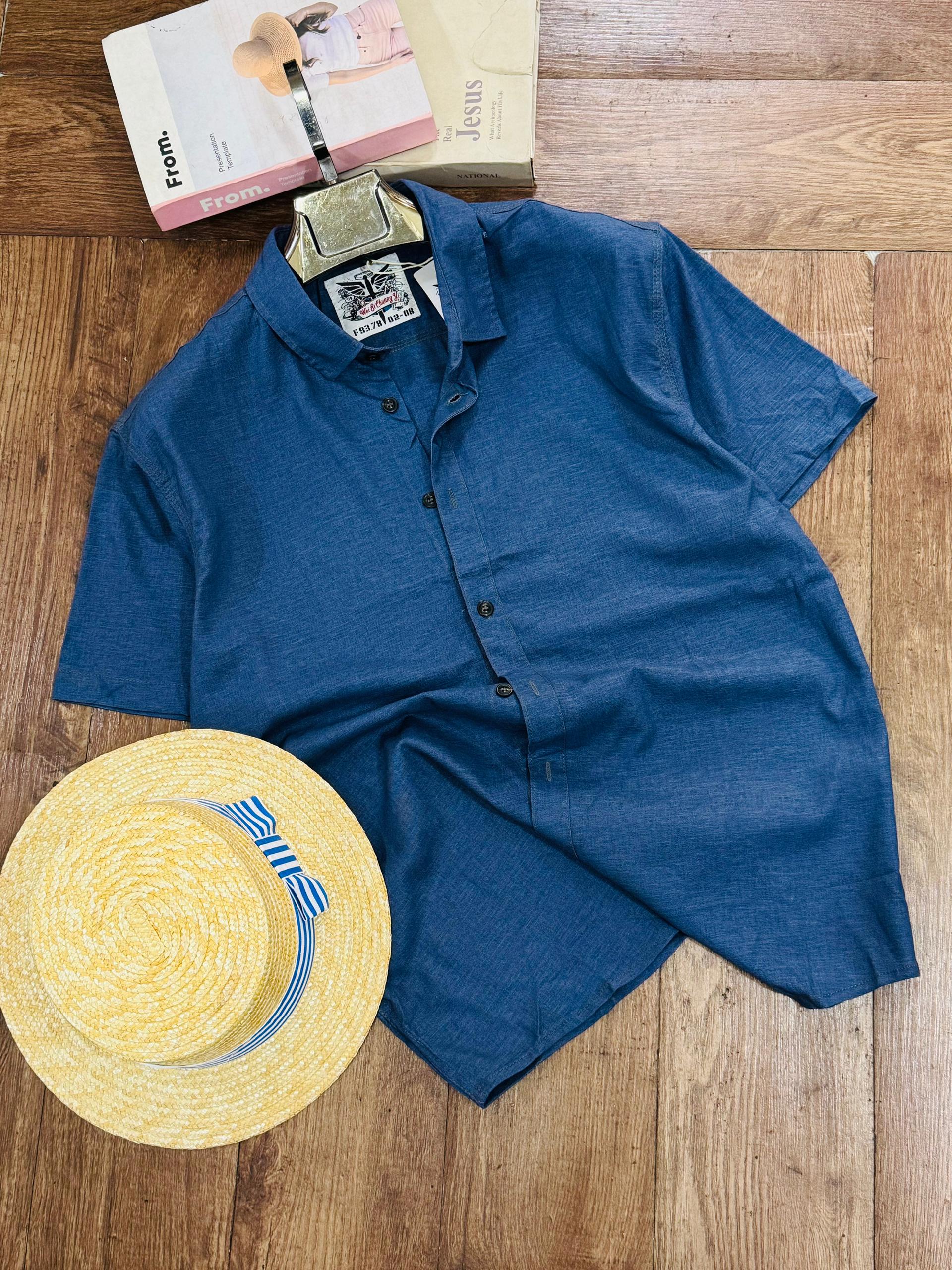 A blue short-sleeve button-up shirt laid flat on a wooden floor, styled with a yellow straw hat and two books.