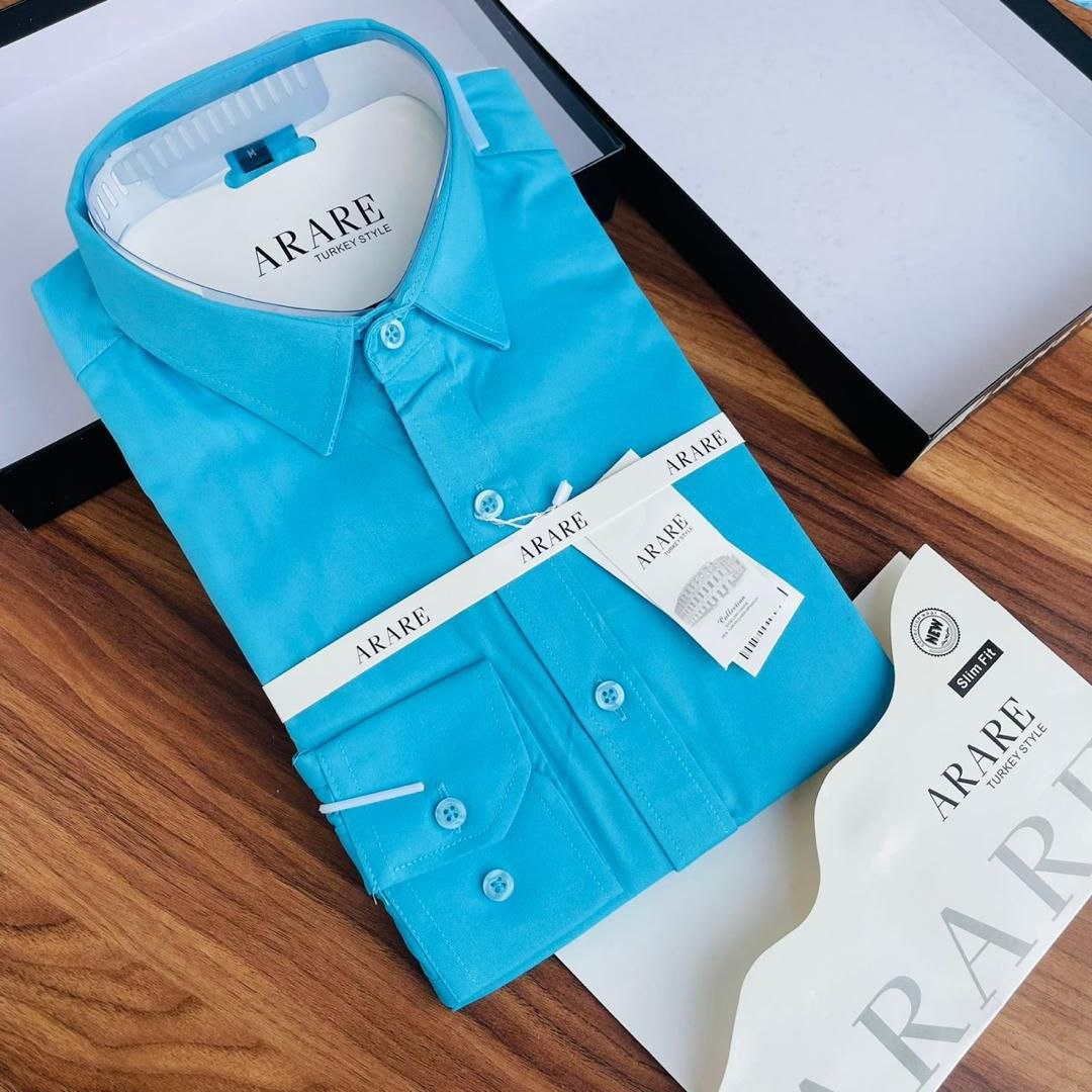 A folded bright turquoise blue ARARE men's dress shirt displayed on a wooden surface with its branded box, hangtag, and paper insert visible.