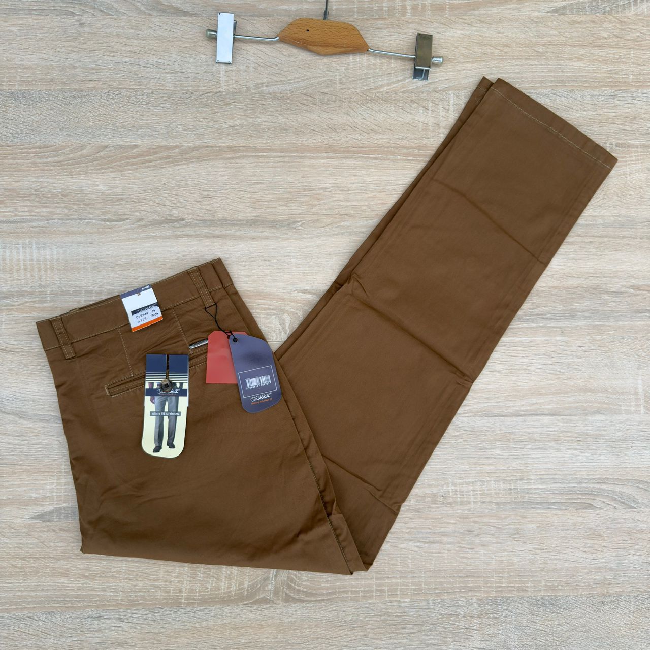 A pair of brown chino pants laid flat on a light wood floor, with retail tags attached near the waistband and a wooden hanger visible at the top of the frame.