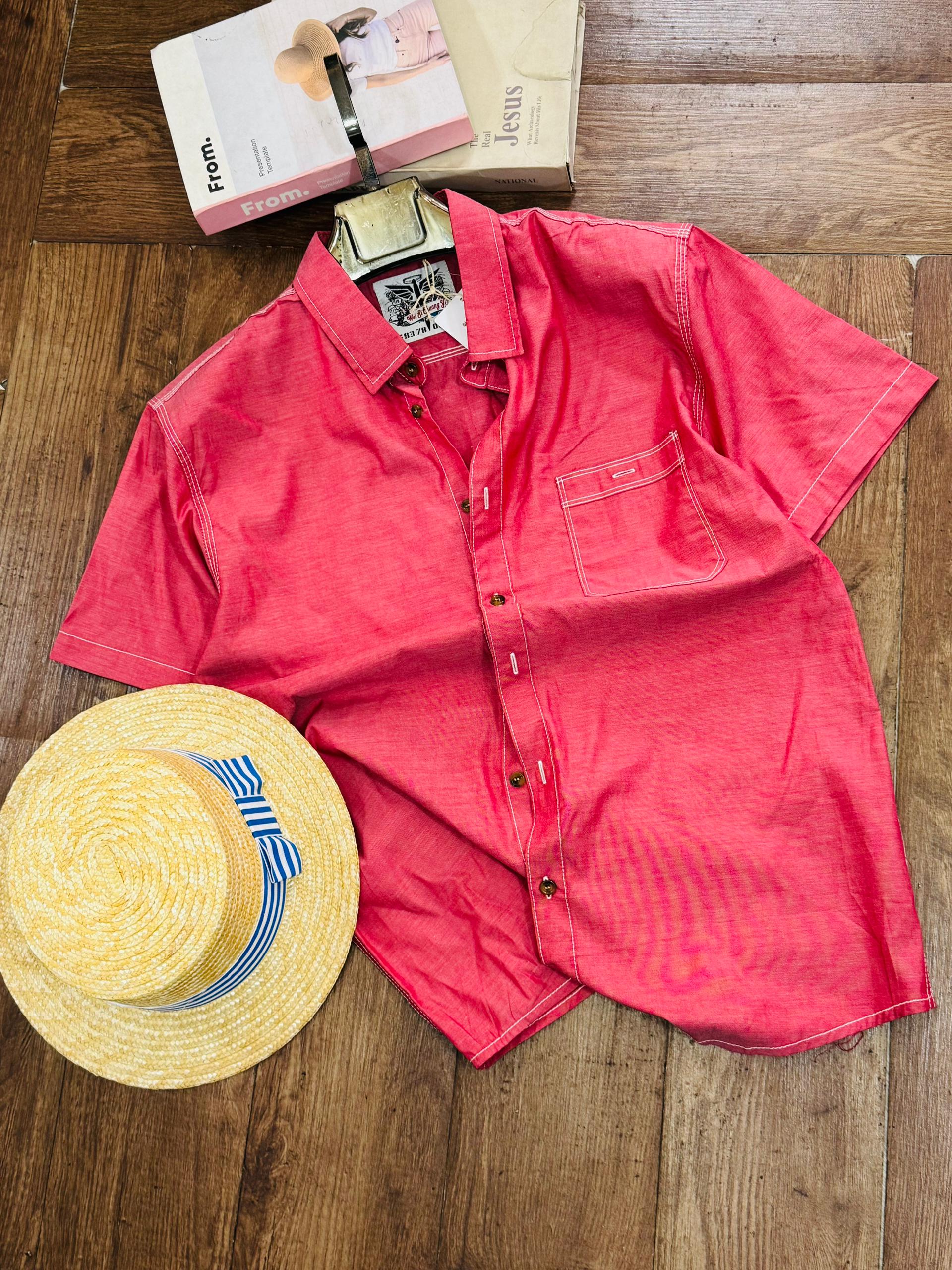 A coral pink short-sleeve button-up shirt laid flat on a wooden floor, styled with a yellow straw hat featuring a blue and white striped band and two product boxes at the top.