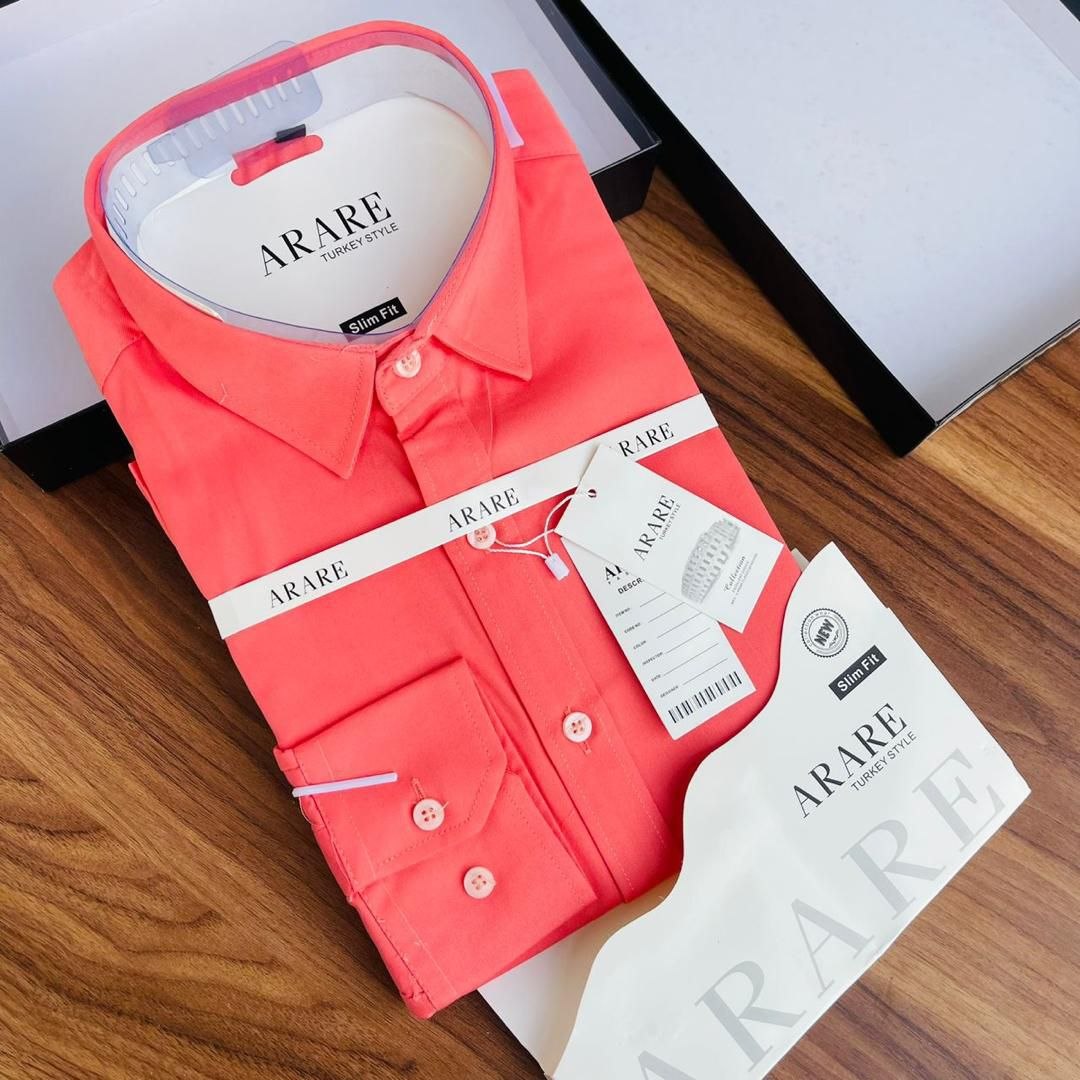 A folded coral pink ARARE men's dress shirt with its white paper packaging, hangtag, and branded ribbon, displayed on a wooden surface.