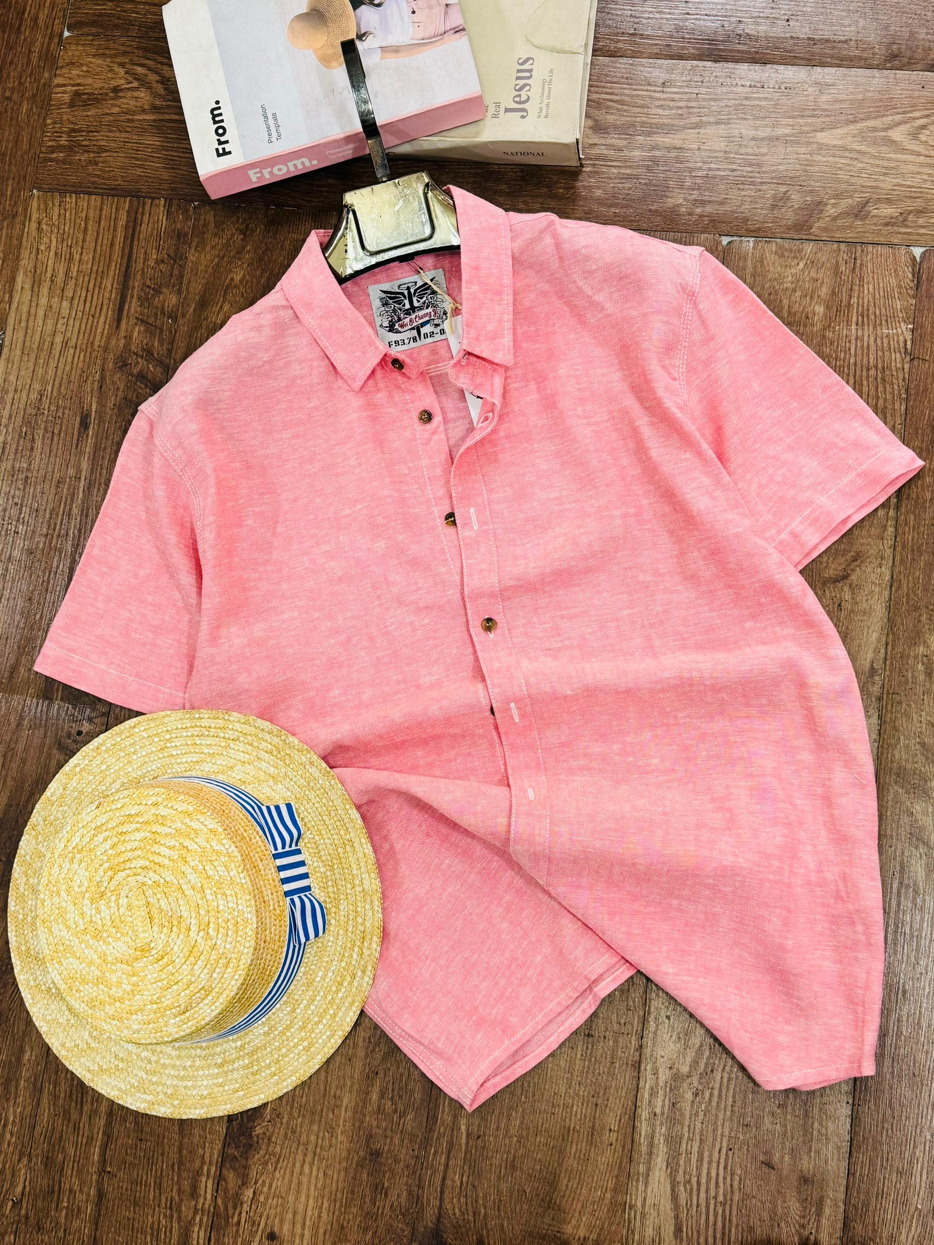 A coral pink short-sleeve button-up shirt laid flat on a wooden floor, styled with a yellow straw hat and a book at the top.