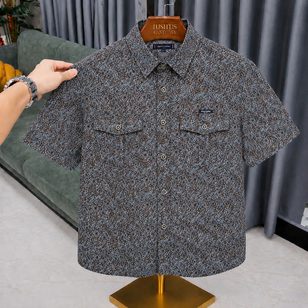 A short-sleeve button-up shirt with a dense blue and gray micro-pattern, displayed on a wooden hanger labeled 'JUSHUS CLOTHING' on a gold stand. A hand is holding the left shoulder of the shirt.