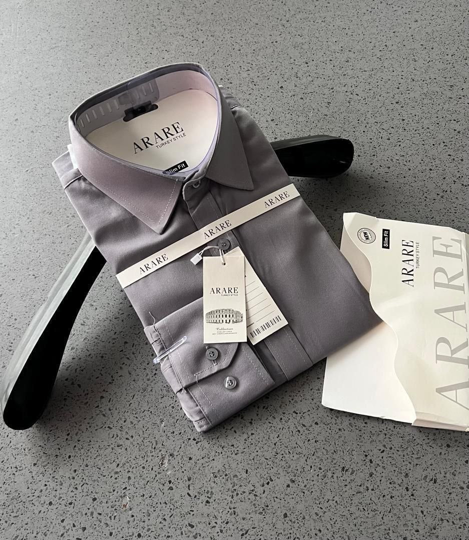 A folded gray long-sleeve dress shirt with a pointed collar and button cuffs, displayed on a black hanger alongside branded ARARE tags and a matching ARARE gift bag on a speckled gray surface.