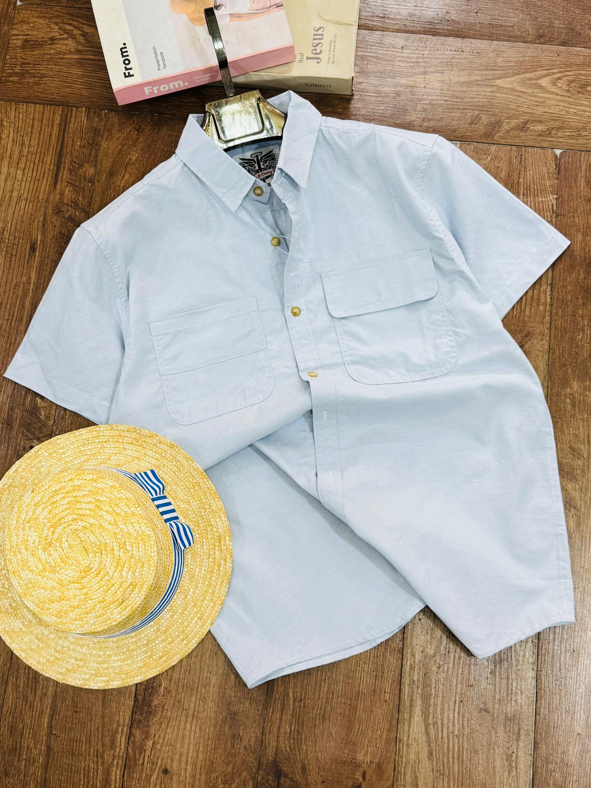 A light blue short-sleeve button-up shirt laid flat on a wooden floor, styled with a yellow straw hat and a book at the top.