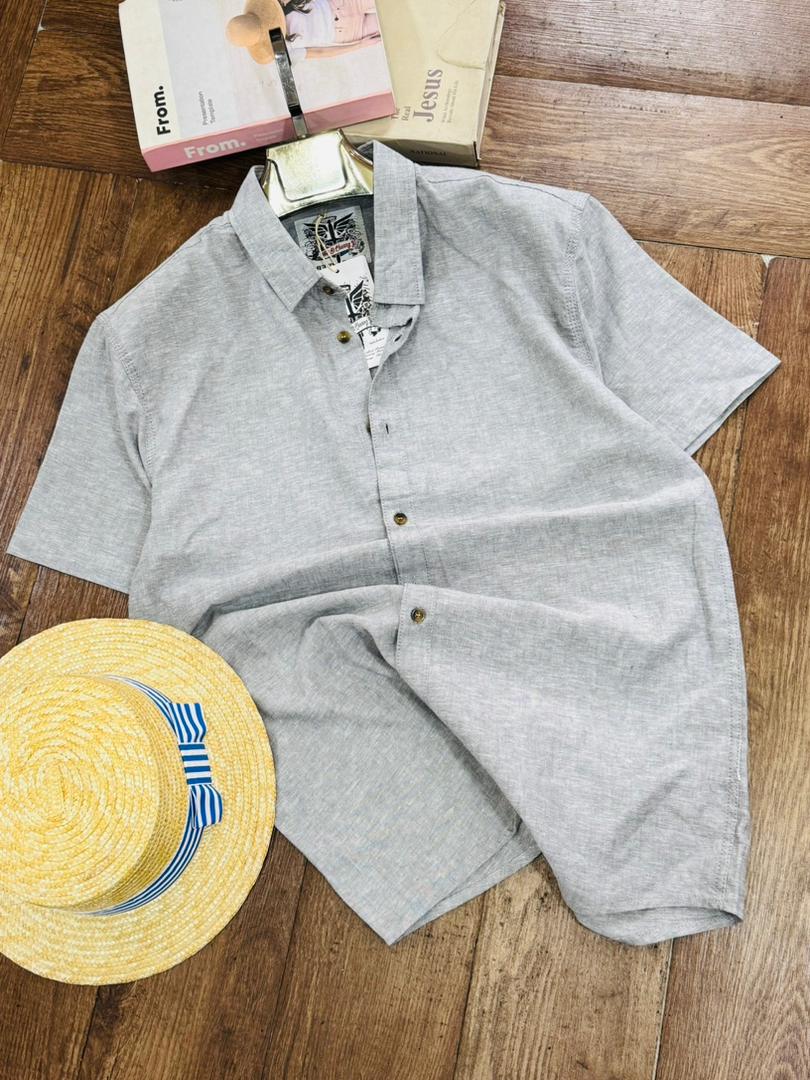 A light gray short-sleeve button-up shirt laid flat on a wooden floor, styled with a yellow straw hat and books.