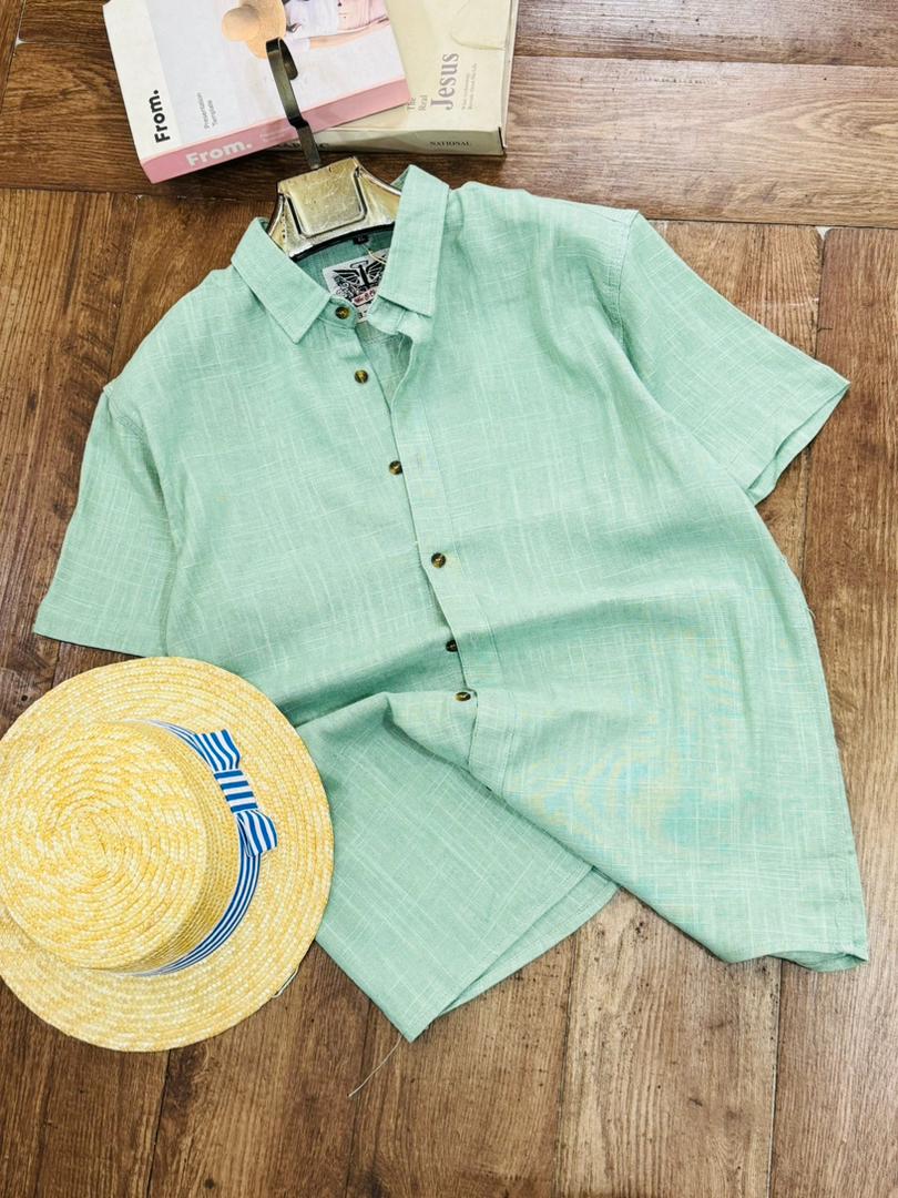 A mint green short-sleeve button-up shirt laid flat on a wooden floor, styled with a yellow straw hat and a book.
