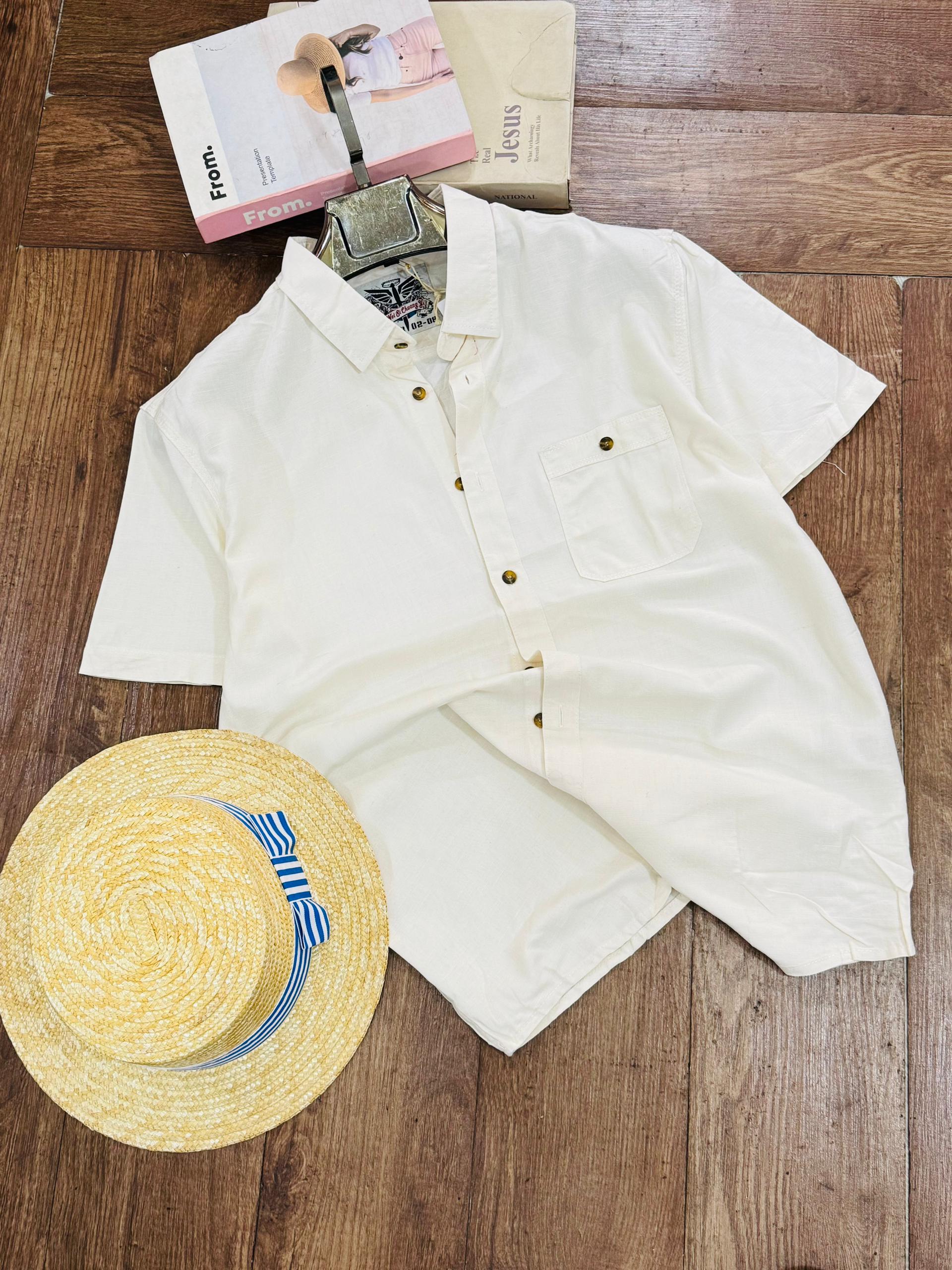 An off-white short-sleeve button-up shirt laid flat on a wooden floor, styled with a yellow straw hat and two magazines.