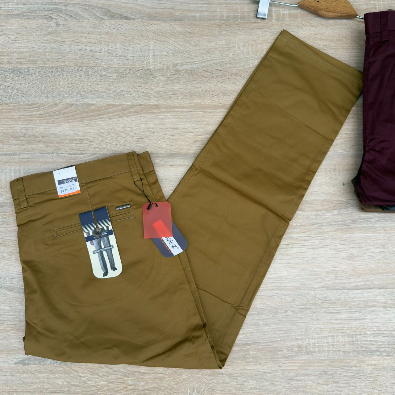 A pair of men's slim-fit chino pants in olive brown, laid flat on a light wood floor. The pants are folded with retail tags attached, including a waistband tag and a pocket label.