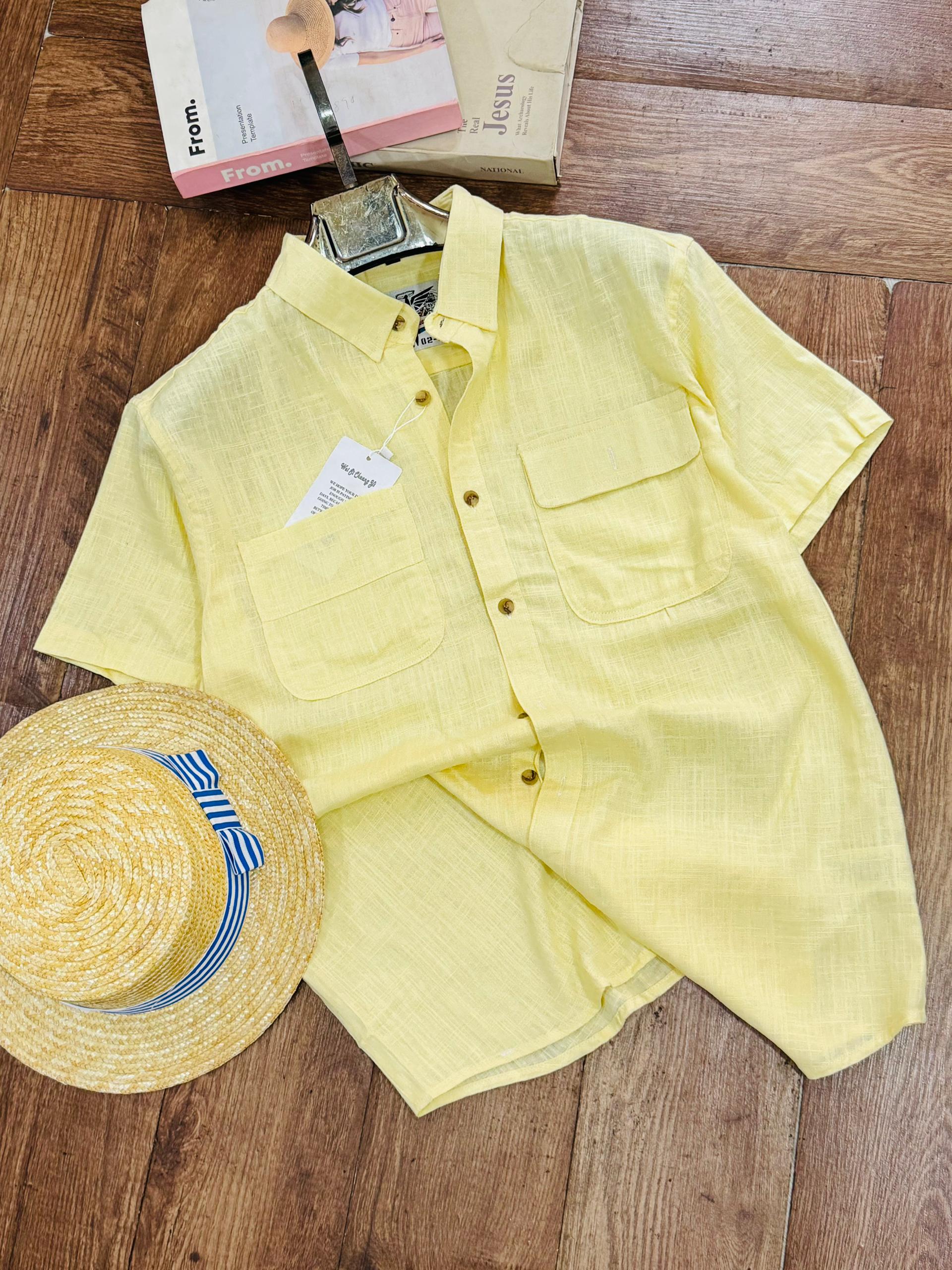 A pale yellow short-sleeve button-up shirt with two chest pockets, laid flat on a wooden floor alongside a straw hat with a blue and white striped band and a small book or magazine.