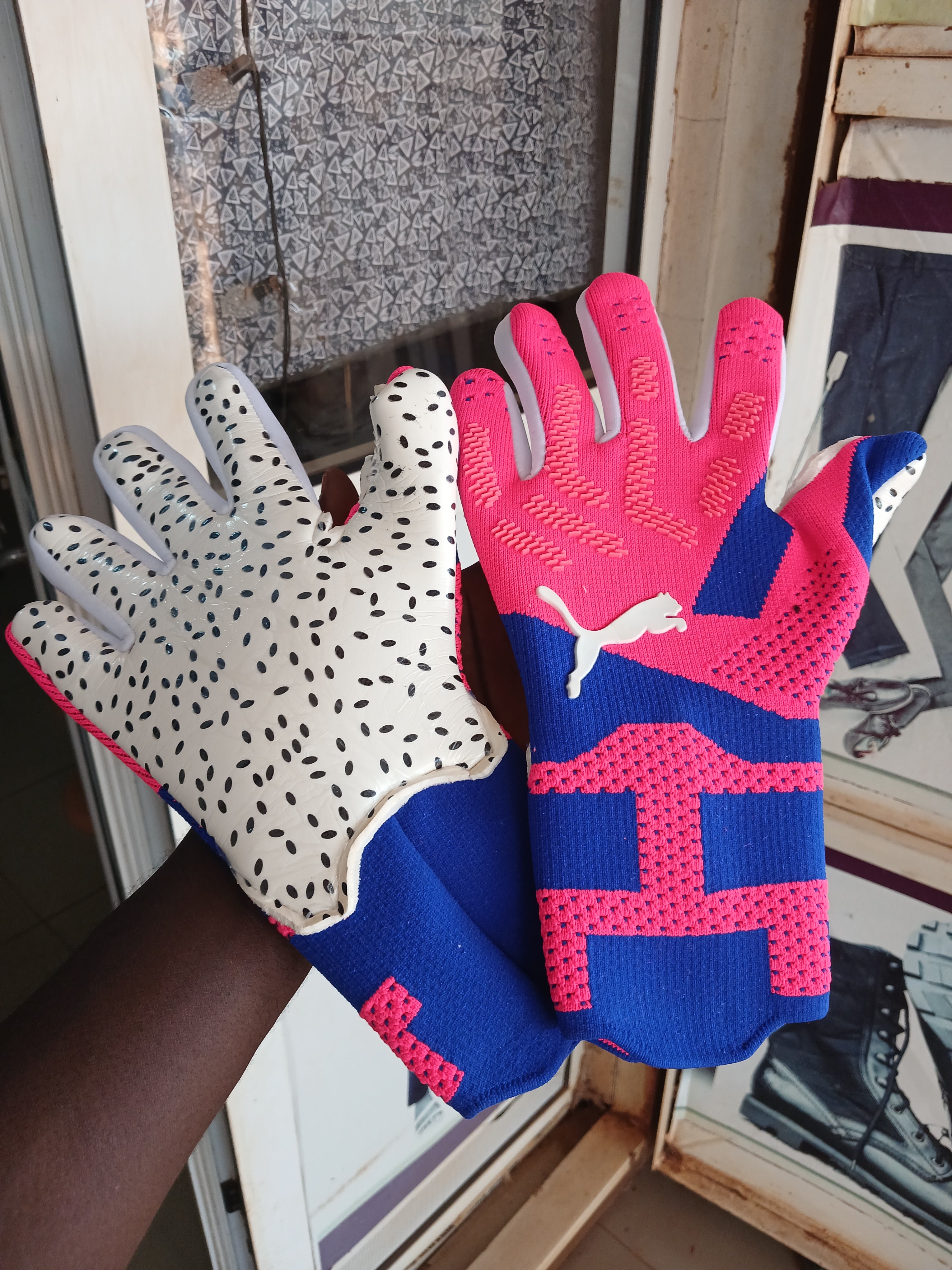 A pair of Puma work gloves held up by a person's hand. One glove is white with black speckles and gray palms, and the other is pink and blue with a white Puma logo on the back.