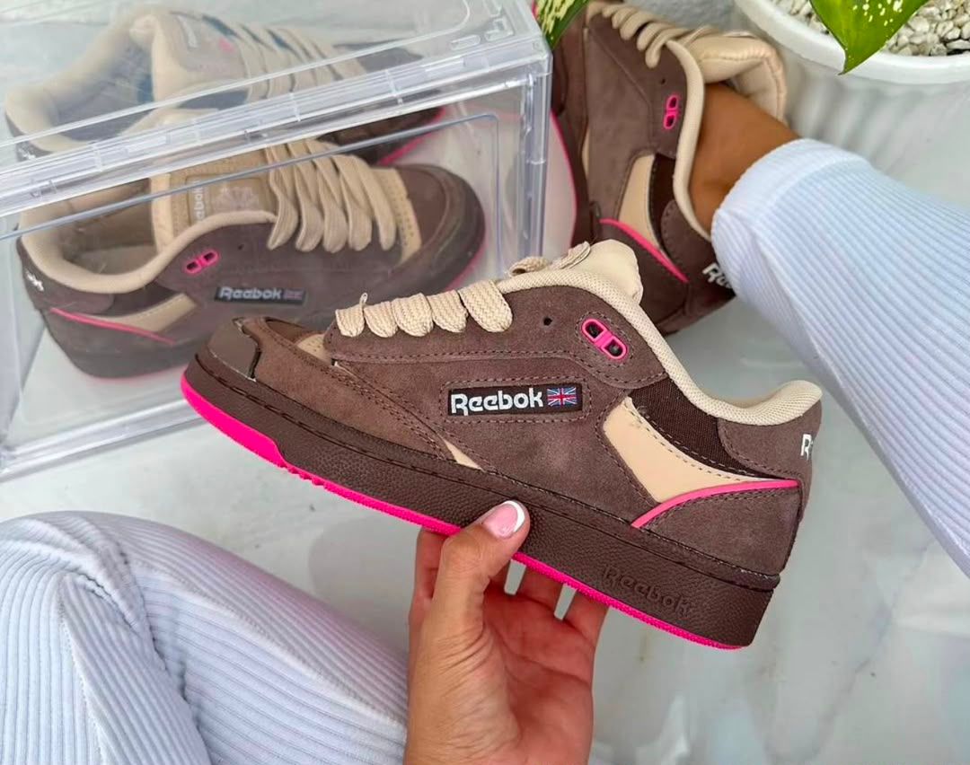 A person holds a brown Reebok low-top sneaker with beige panels and bright pink accents, with other shoes and a clear shoebox visible in the background.