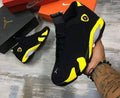 A black Jordan sneaker with bright yellow and gray accents is held by a tattooed hand over a gray wood floor, with another shoe, orange Nike boxes, and a black Jordan box in the background.