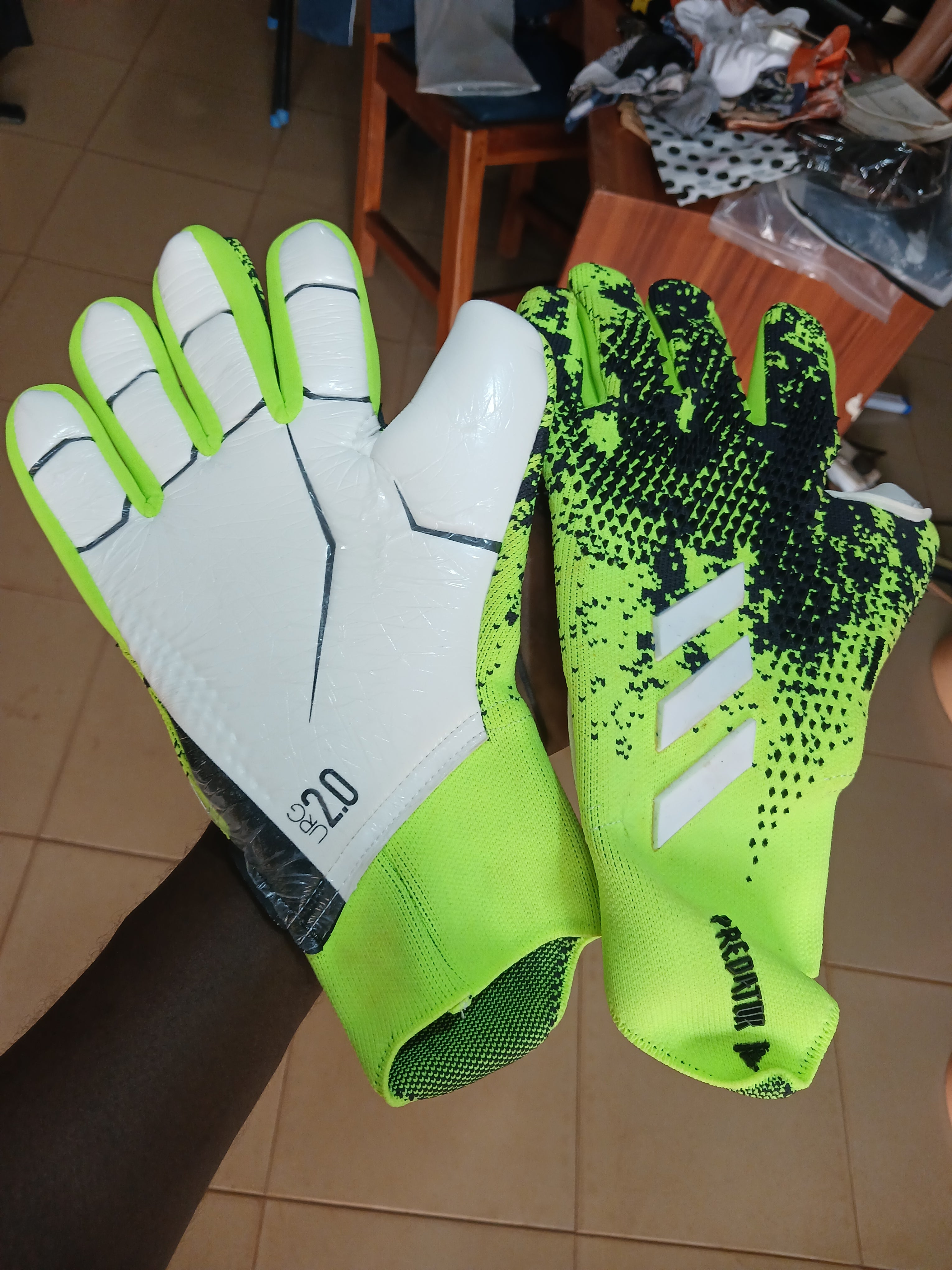 A pair of Adidas Predator soccer gloves held up by a person's hand. The gloves feature a neon green and black speckled pattern on one glove, and a white and neon green design on the other, with the Adidas three-stripe logo and 'PREDATOR' branding visible.